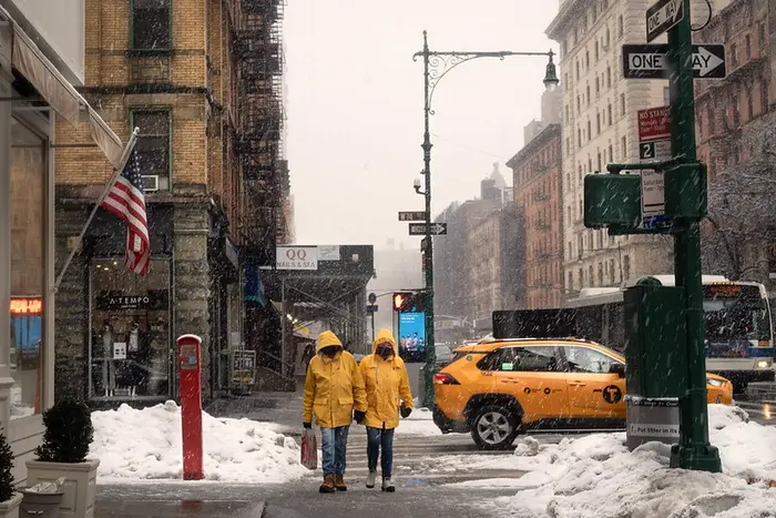 Two people in yellow snow jackets walk past a yellow cab in snowfall in NYC.